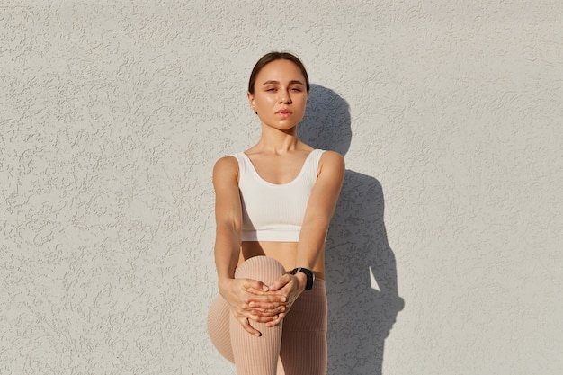 Prise de vue en plein air de jeunes vêtus d'un haut blanc, debout sur une jambe, touchant le genou avec les mains, regardant la caméra avec une expression faciale sérieuse.