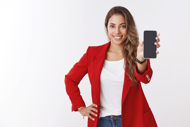 Prise de vue en studio agréable souriante heureuse fille caucasienne blonde souriante dents blanches montrant l'écran d'affichage du smartphone, debout confiant présentant l'application, suggérant d'utiliser le mur blanc de l'application