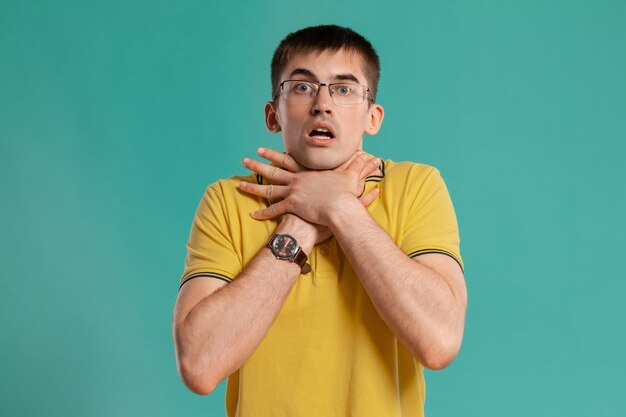 Prise de vue en studio d'un beau jeune homme dans un t-shirt décontracté jaune, des lunettes et des montres noires s'étouffant en posant sur un fond bleu. Coupe de cheveux élégante. Concept d'émotions sincères. Copiez l'espace.