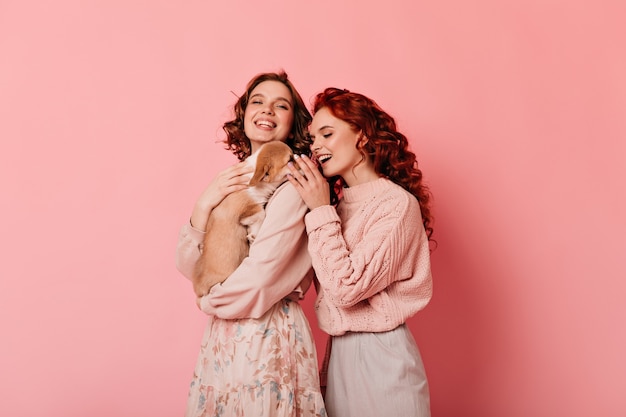 Prise de vue en studio de deux amis avec un chien. Filles bouclées jouant avec chiot sur fond rose.