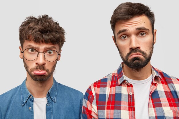 Prise de vue en studio de deux beaux hommes portés se sentent insultés, courbent les lèvres, ont des expressions déplacées, sont offensés à cause de quelque chose, posent contre un mur blanc