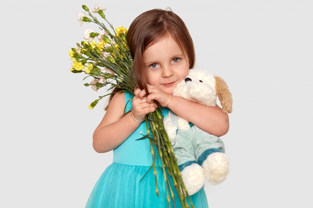 Prise de vue en studio d'un enfant attrayant porte étroitement des fleurs et une peluche, vêtue d'une robe bleue gonflée. Concept d'enfance.
