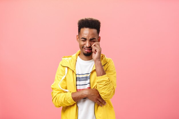 Prise de vue en studio d'un homme timide afro-américain triste et idiot en veste jaune à la mode pleurant le cœur en fouettant les larmes des yeux et se sentant seul après avoir été rejeté par son ex-petite amie sur un mur rose