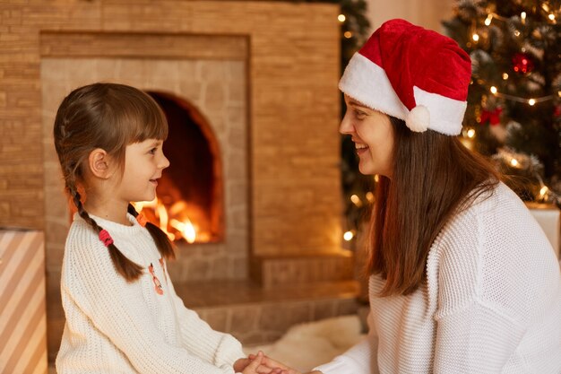 Prise de vue en studio intérieur d'une femme aux cheveux noirs portant un chapeau de père Noël avec sa petite fille se tenant la main et se regardant avec beaucoup d'amour. posant dans un salon festif près de la cheminée.