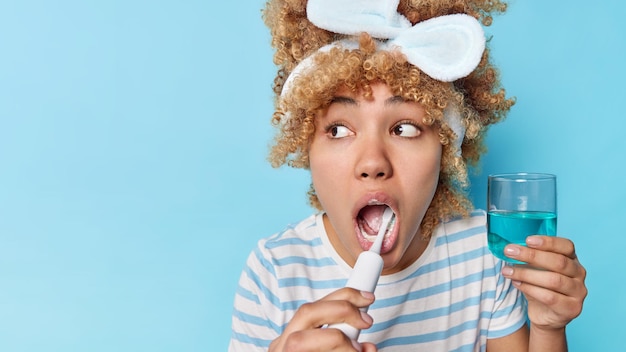 Prise de vue en studio d'une jeune femme aux cheveux bouclés surprise regarde ailleurs se brosse les dents prend soin de l'hygiène bucco-dentaire utilise une brosse électrique tient des poses de rince-bouche sur fond bleu espace vide pour votre texte