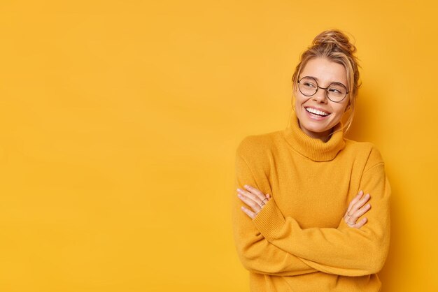 Prise de vue en studio d'une jeune femme positive aux cheveux blonds peignés garde les bras croisés se sent satisfaite regarde quelque chose porte un pull et des lunettes isolées sur fond jaune espace vide pour votre texte