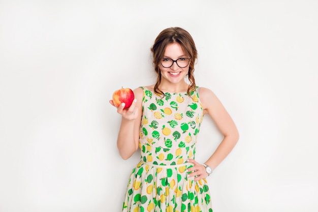 Prise de vue en studio d'une jeune fille brune souriante souriant largement. La fille porte des lunettes noires et une robe jaune et verte. Elle tient une pomme rouge dans sa main droite.