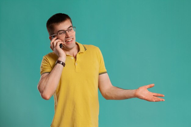 Prise de vue en studio d'un jeune homme élégant dans un t-shirt décontracté jaune, des lunettes et des montres noires parlant par téléphone tout en posant sur un fond bleu. Coupe de cheveux élégante. Concept d'émotions sincères. Copiez l'espace.