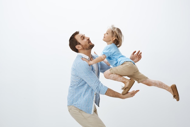 Prise de vue en studio d'un père attrapant un fils blond mignon qui saute de table en jouant et en s'amusant en s'occupant d'un garçon pendant que sa mère au travail marche sur une aire de jeux ensemble en famille sur un mur gris