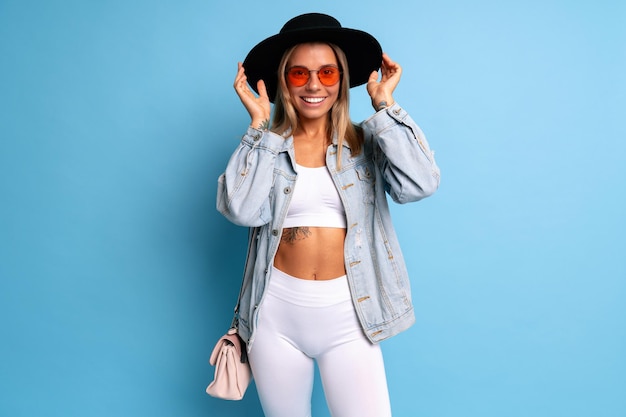 Prise de vue en studio positive à la mode d'une femme hipsyer b;onde vêtue d'un costume sportif blanc, d'une veste en jean et d'un chapeau fedora noir.