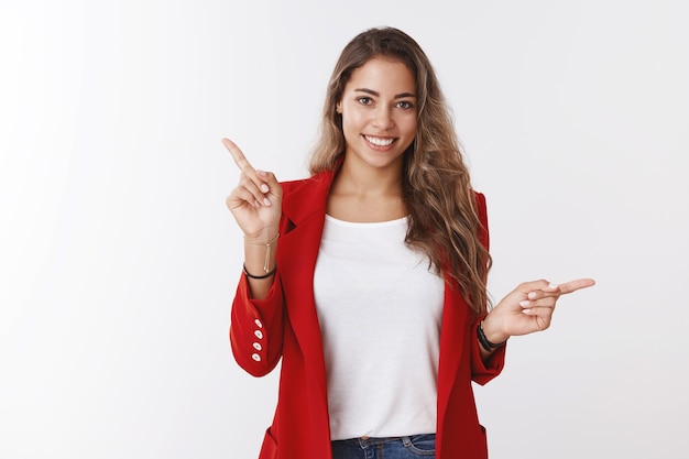 Prise de vue en studio séduisante sympathique souriante heureuse caucasienne de 25 ans portant une veste rouge pointant latéralement dans différentes directions souriante présentant des choix souriant suggérant des marchandises, mur blanc