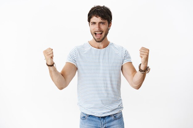 Prise de vue à la taille d'un bel homme souriant et souriant aux yeux bleus et aux poils en t-shirt rayé criant et levant les poings serrés comme triomphant, étant fan et équipe de soutien sur un mur blanc