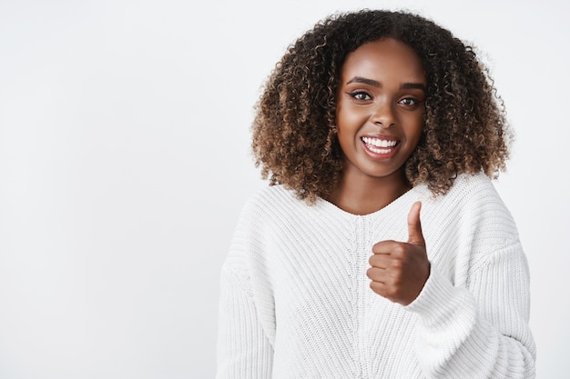 Prise de vue à la taille d'une femme montrant le pouce vers le haut et souriant joyeusement recommandant le produit, aimant la superbe tenue d'un ami comme donnant une opinion positive personnelle, souriant largement sur un mur blanc