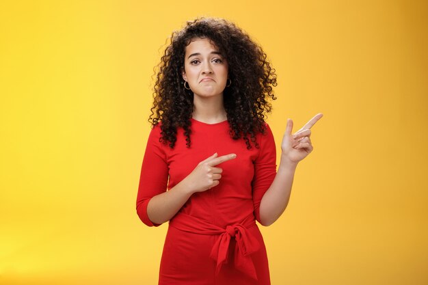 Prise de vue à la taille d'une fille mécontente bouleversée avec des cheveux bouclés étant dans les décharges pointant vers le coin supérieur droit avec un sourire sombre et déçu, ressentant du regret, de la tristesse debout comme un perdant sur un mur jaune.