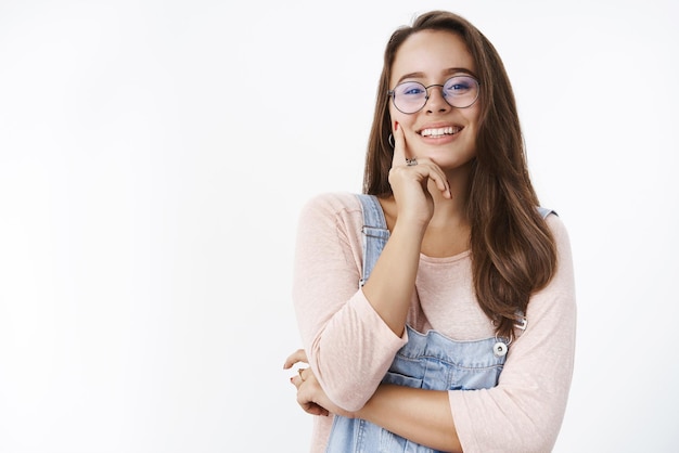 Prise de vue à la taille d'une jeune femme sensuelle et heureuse des années 20 dans des lunettes et une salopette en jean touchant la joue avec l'index souriant largement avec un regard confiant posant satisfait sur fond gris