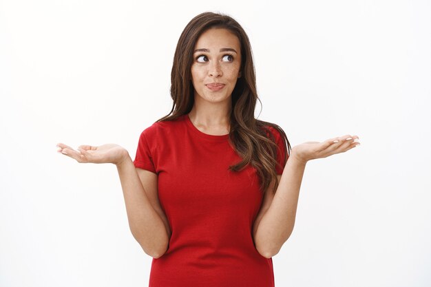 Prise de vue à la taille mignonne femme brune idiote en t-shirt décontracté rouge, haussant les mains levant les mains interrogées, souriant et détournant le regard en faisant un choix, en réfléchissant entre les variantes, en se tenant debout sur un mur blanc