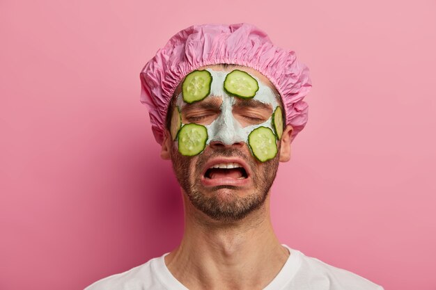 Procédure de beauté, cosmétologie et concept de soins de la peau. Un homme insatisfait mélancolique pleure d'émotions négatives, porte un bonnet de bain, des concombres sur le visage