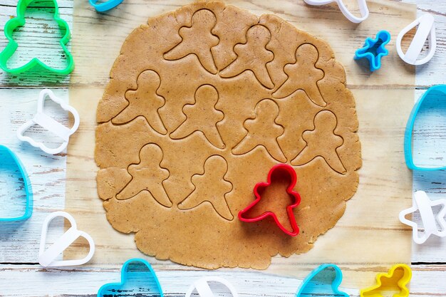 Processus de traitement des biscuits de pain d'épice, utilisez le moule de pain d'épice de coupe de pain d'épice sur du papier sulfurisé autour de découpeurs de biscuits colorés sur une table en bois blanche. vue de dessus