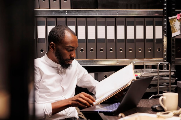 Procureur afro-américain lisant un dossier, se préparant à une affaire au bureau de police avec des documents d'archives. Professionnel de l'application de la loi analysant le dossier d'expertise médico-légale la nuit