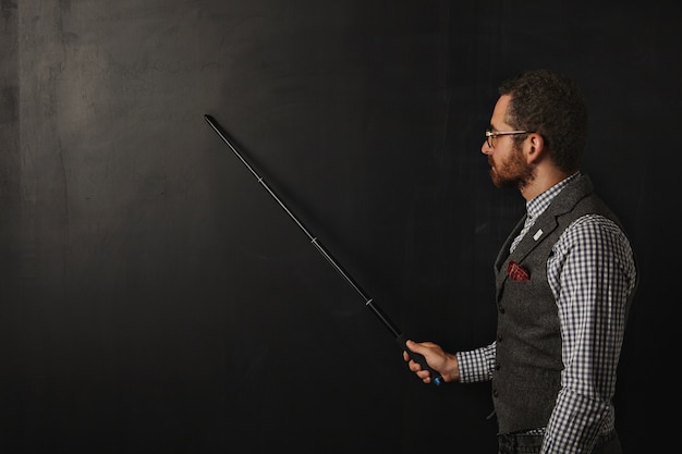 Professeur barbu sérieux en chemise à carreaux et gilet en tweed, portant des lunettes, montre quelque chose sur le tableau noir de l'école avec son pointeur pliant