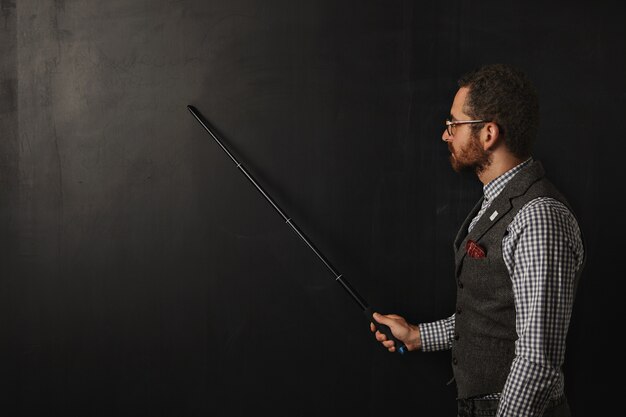 Professeur barbu sérieux en chemise à carreaux et gilet en tweed, portant des lunettes, montre quelque chose sur le tableau noir de l'école avec son pointeur pliant