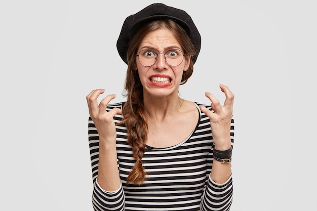 Un Professeur De Français Irrité Serre Les Dents Et Fait Des Gestes Avec Colère, Regarde Avec Impatience, A Une Expression Faciale Négative, Porte Un Béret, Pose Sur Un Mur Blanc