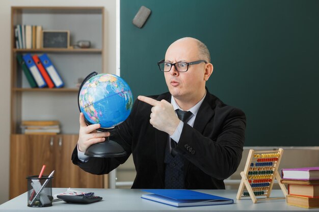 Professeur d'homme portant des lunettes assis avec globe au bureau de l'école devant le tableau noir dans la classe expliquant la leçon à la surprise