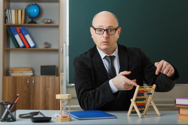 Professeur d'homme portant des lunettes avec registre de classe assis au bureau de l'école devant le tableau noir dans la salle de classe à l'aide d'abaque à la confusion