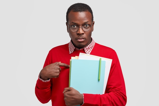 Un professeur indigné se montre du doigt, pose des questions sur ses fonctions, porte un manuel, porte un pull rouge décontracté, va organiser un séminaire pour les scientifiques isolé sur un mur blanc