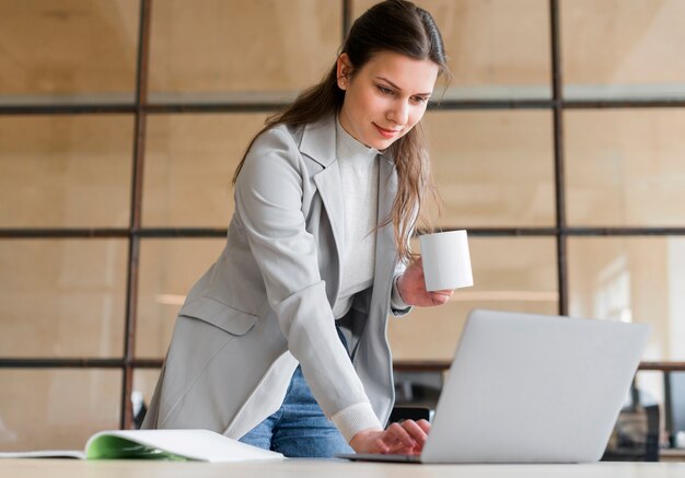 Professional jeune femme souriante tenant une tasse de café blanc travaillant sur ordinateur portable