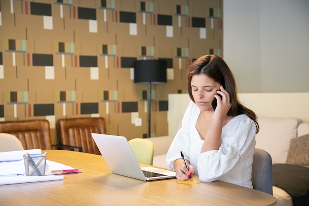 Professionnel de la création ciblé discutant du projet de conception avec le client au téléphone, assis à table avec un ordinateur portable et des plans et des notes d'écriture