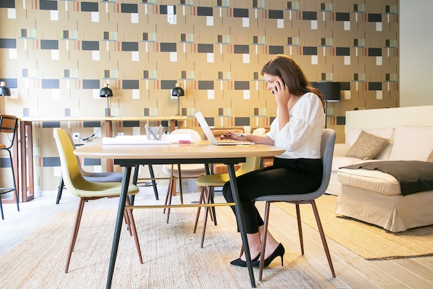 Professionnel de la création confiant discutant du projet avec le client au téléphone, assis à table avec un ordinateur portable et des plans et dactylographie