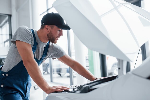 Professionnel sur le travail. Homme en uniforme bleu et chapeau noir réparant une automobile endommagée