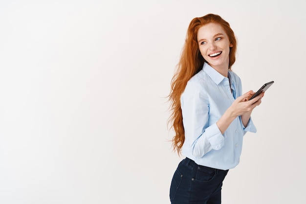 Profil d'une belle gestionnaire aux longs cheveux roux utilisant un téléphone portable, tournez la tête à gauche et souriant à l'espace de copie, debout avec un smartphone contre un mur blanc