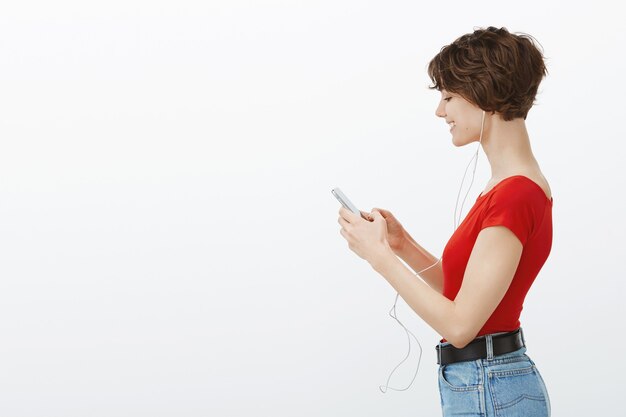 Profil de belle jeune femme regardant smartphone et écouter de la musique dans des écouteurs, bénéficiant d'un podcast
