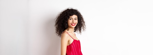 Photo gratuite profil d'une femme aux cheveux bouclés à la mode en robe rouge tourner la tête à l'appareil photo et sourire confiant stan