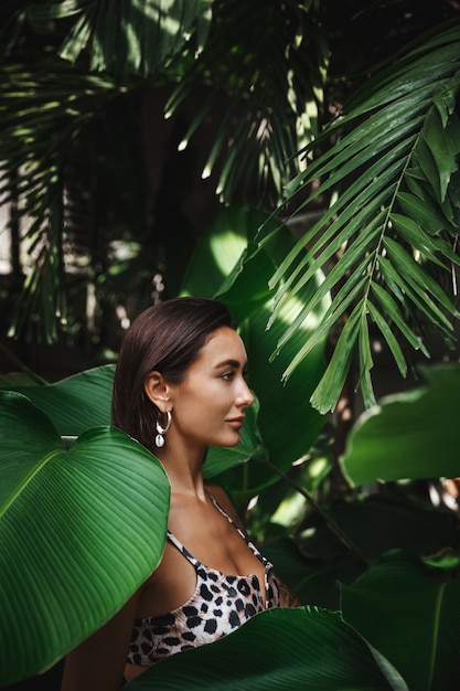 Profil de femme bronzée en bikini, portant des boucles d'oreilles, debout dans les feuilles tropicales de la jungle et les palmiers.