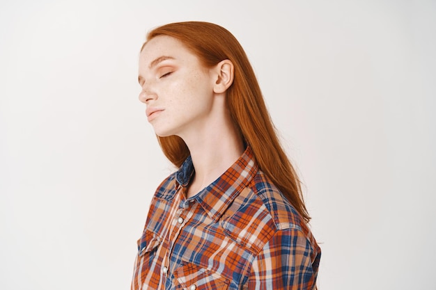 Profil d'une femme tendre et détendue aux cheveux longs roux, aux yeux fermés et au cou incliné, debout sur un mur blanc