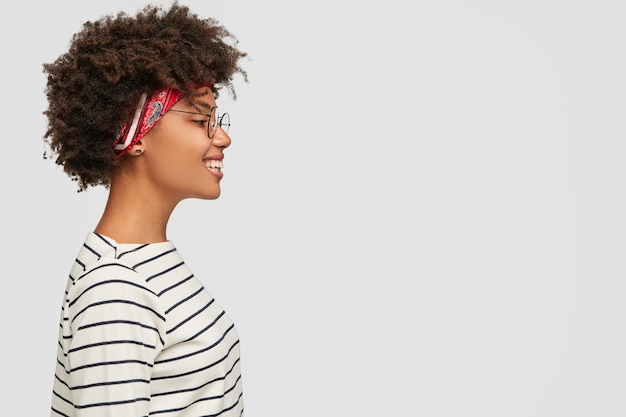 Photo gratuite profil de fille heureuse à la peau sombre porte des vêtements à rayures, un bandeau, des lunettes