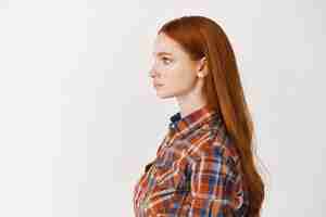 Photo gratuite profil d'une jeune femme aux longs cheveux roux en bonne santé et à la peau pâle, regardant à gauche avec un visage sérieux, debout sur un mur blanc