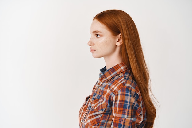 Profil d'une jeune femme aux longs cheveux roux en bonne santé et à la peau pâle, regardant à gauche avec un visage sérieux, debout sur un mur blanc