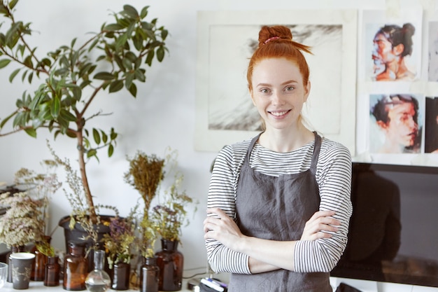 Photo gratuite profitant du processus de création. séduisante jeune artisan, artiste ou designer au gingembre avec un nœud de cheveux, se sentant heureuse et enthousiaste à l'idée de créer quelque chose de beau et de nouveau dans son studio