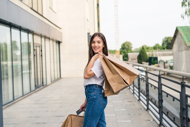 Profiter des achats de jour. Sur toute la longueur de la jeune femme tenant des sacs à provisions et souriant en marchant le long de la rue