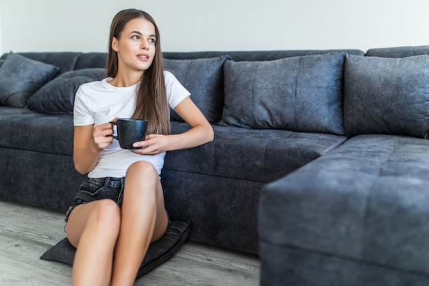 Profiter du café à la maison. Belle jeune femme souriante assise sur le tapis à la maison et tenant une tasse de café