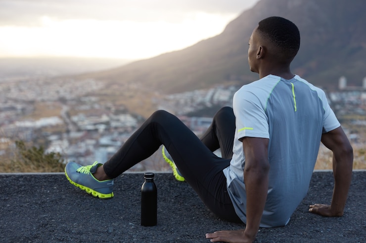 Photo gratuit profondément dans ses pensées, un homme noir porte des vêtements de sport décontractés, se sent détendu au sommet, pose devant la vue panoramique sur la montagne, garde les mains sur l'asphalte, s'entraîne en plein air, fatigue et manque de force