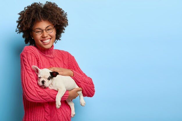 Le propriétaire d'un animal à la peau foncée attentionné tient un petit chiot, aime les animaux de compagnie, porte des lunettes et un pull rose, sourit joyeusement