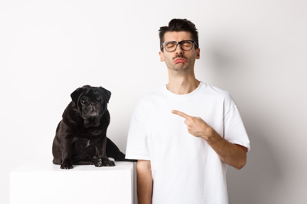 Propriétaire d'animal triste et sombre pointant vers son chien carlin noir et sanglotant, debout upster sur fond blanc