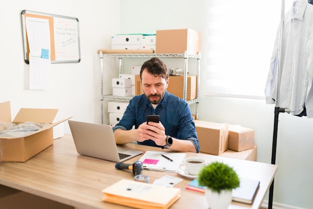 Le propriétaire d'une entreprise latine envoie des SMS sur le smartphone et met à jour ses médias sociaux professionnels. Jeune entrepreneur avec une boutique en ligne