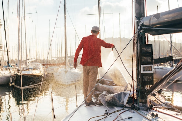 Propriétaire de voilier ou plaisancier utilise le tuyau pour laver l'eau salée du pont de yachr lorsqu'il est amarré ou stationné dans la marina au coucher du soleil