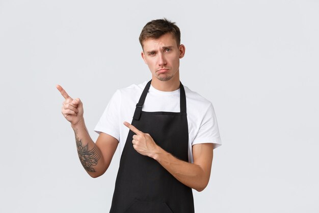 Propriétaires de petites entreprises, café et concept de personnel. Bouleversé et sombre, mignon barista faisant un visage triste et pointant du doigt le coin supérieur gauche déçu, se plaignant ou racontant de mauvaises nouvelles